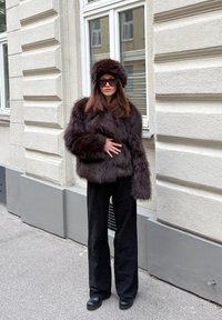 Jachetă maro din blană artificială cu o pălărie asortată, croială largă, purtată cu pantaloni negri cu picioare largi și ochelari de soare rotunzi, pe fundalul unui zid texturat.