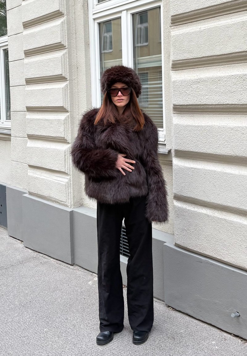 Veste en fausse fourrure marron avec un chapeau assorti, coupe oversize, portée avec un pantalon noir à jambes larges et des lunettes de soleil rondes, devant un mur texturé.