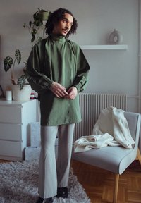 Persoon met krullend haar staat binnen in een groene langslipper en lichtgrijze broek, naast een stoel met een witte gebreide deken.