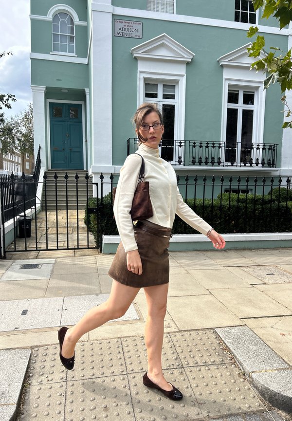 Model trägt einen cremefarbenen, geknöpften Rollkragenpullover und einen braunen Wildleder-Minirock, hält eine braune Tasche und geht über eine Kopfsteinpflasterstraße.