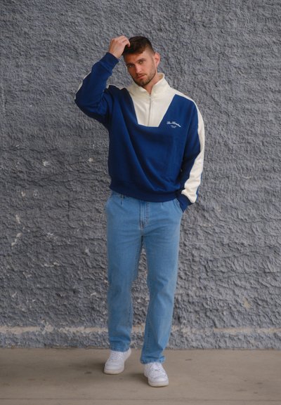 Hombre con sudadera azul y blanca con cremallera, jeans azul claro y zapatillas blancas, de pie contra una pared gris texturizada, tocándose el cabello.