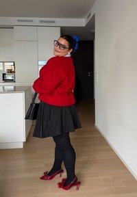 Crvena pletena džemper, crna naborana suknja, crne hulahopke i crvene salonke s remenčićem i mašnama. Plavi ukras za kosu. Moderna kuhinja u pozadini.