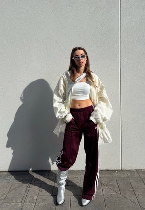 Mujer con top corto blanco, chaqueta ligera oversized, pantalones deportivos burdeos, botas plateadas y gafas de sol, de pie contra una pared blanca con las manos en los bolsillos.