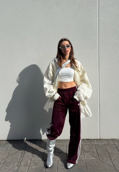 Mujer con top corto blanco, chaqueta ligera oversized, pantalones deportivos burdeos, botas plateadas y gafas de sol, de pie contra una pared blanca con las manos en los bolsillos.
