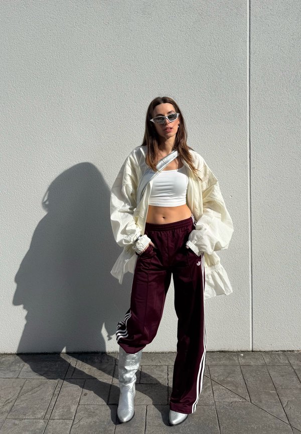 Femme portant un crop top blanc, une veste légère surdimensionnée, un pantalon de jogging bordeaux, des bottes argentées et des lunettes de soleil, debout contre un mur blanc les mains dans les poches.
