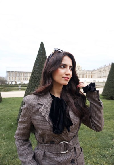 Mujer con abrigo de cuadros, lazo negro y gafas de sol en la cabeza, de pie en un jardín con árboles cónicos recortados y un edificio histórico al fondo.
