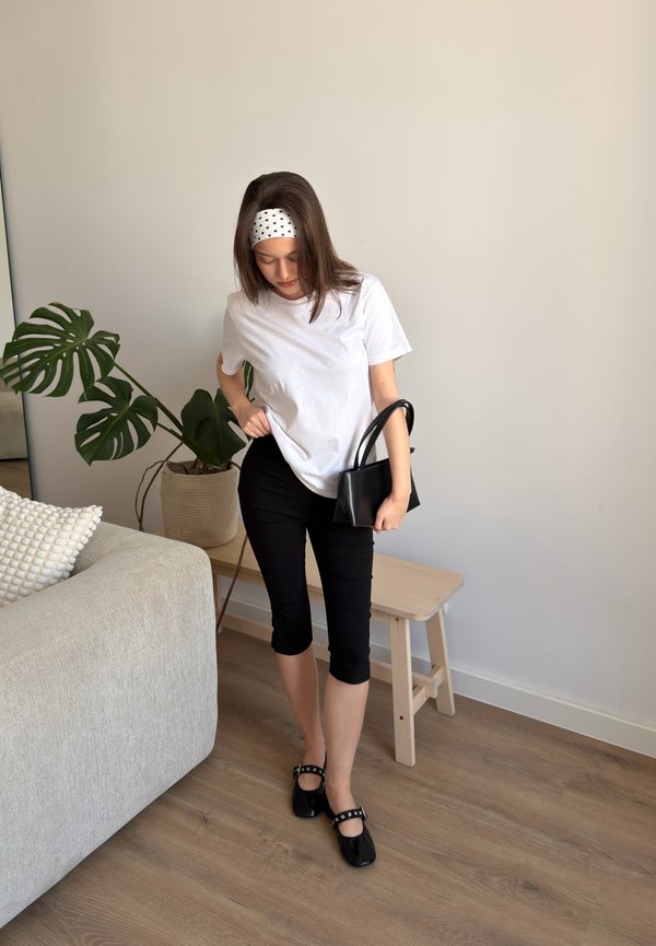 Femme portant un t-shirt blanc, un pantalon capri noir, des chaussures noires et un bandeau à pois, debout à l'intérieur tenant un sac à main noir près d'un banc et d'une plante.