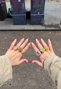 Kaks kätt, millel on mitmed sõrmused: kuldsed rõngad, roosa käekell, hõbedane sõrmus punase kiviga ning punaste otstega hoolitsetud küüned.