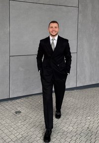 Mand i sort dobbeltradet jakkesæt og slips går på flisebelagt fortov med hænderne i lommerne foran en grå flisevæg.