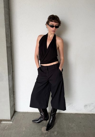 Persona con top negro sin mangas con escote cruzado, pantalones anchos recortados, botas negras puntiagudas y gafas de sol oscuras, de pie contra una pared blanca.
