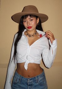 Giovane donna con un cappello a tesa larga marrone, rossetto rosso, camicia bianca annodata, jeans blu e collana a forma di cuore, che mostra una fascia rossa sotto la camicia.