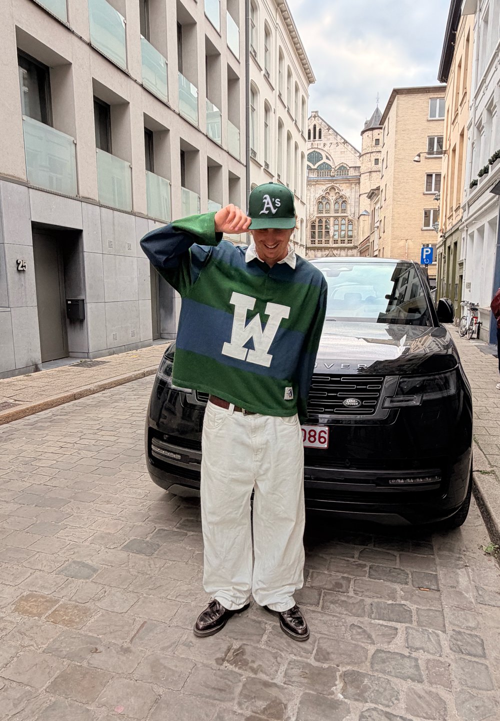Zeleno-navy pruhovaný ragbyový dres s bielym golierom, s veľkým bielym logom "W". Bielé oversize nohavice a čierne topánky. Čierne SUV v pozadí.