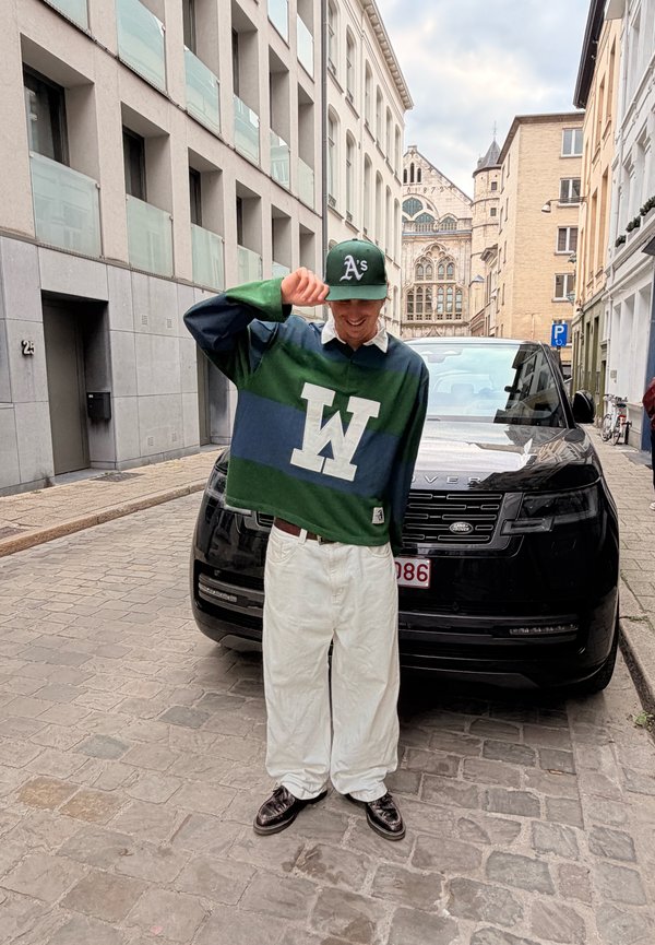 Verde e azul listrado polo de rugby com colarinho branco, apresentando um grande logotipo branco "W". Calças brancas oversized e sapatos pretos. SUV preto ao fundo.