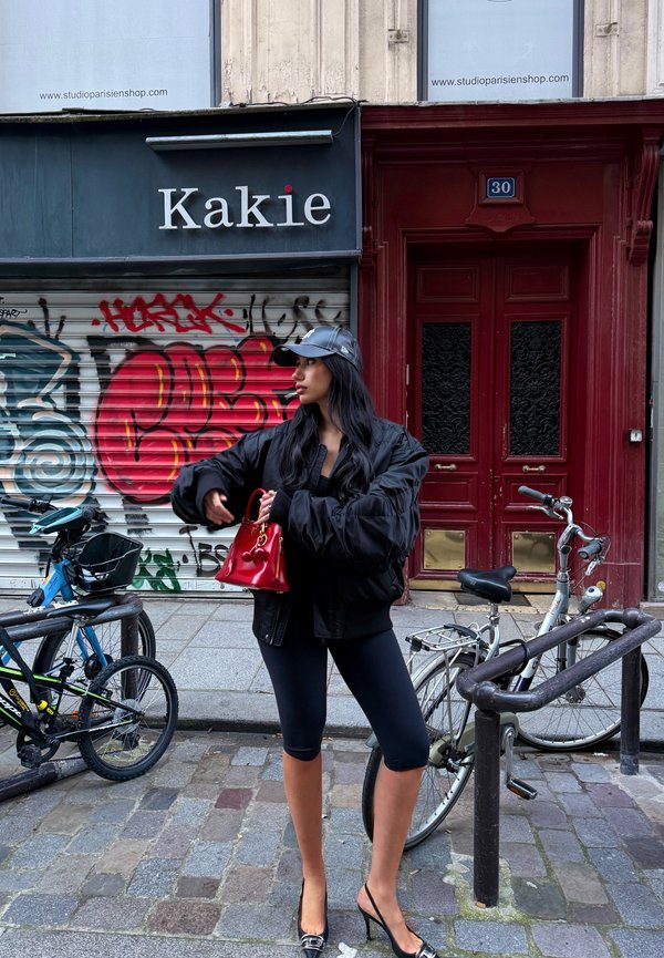 Svart oversize-jacka, tajta svarta leggings, höga klackar. Håller en liten röd handväska med dekorativa detaljer. Graffiti i bakgrunden, cyklar i närheten.