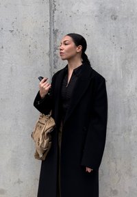 Manteau en laine noire par-dessus un chemisier foncé, tenant un sac en cuir beige. Couleurs lisses et unies avec des motifs minimaux. Fond en béton.