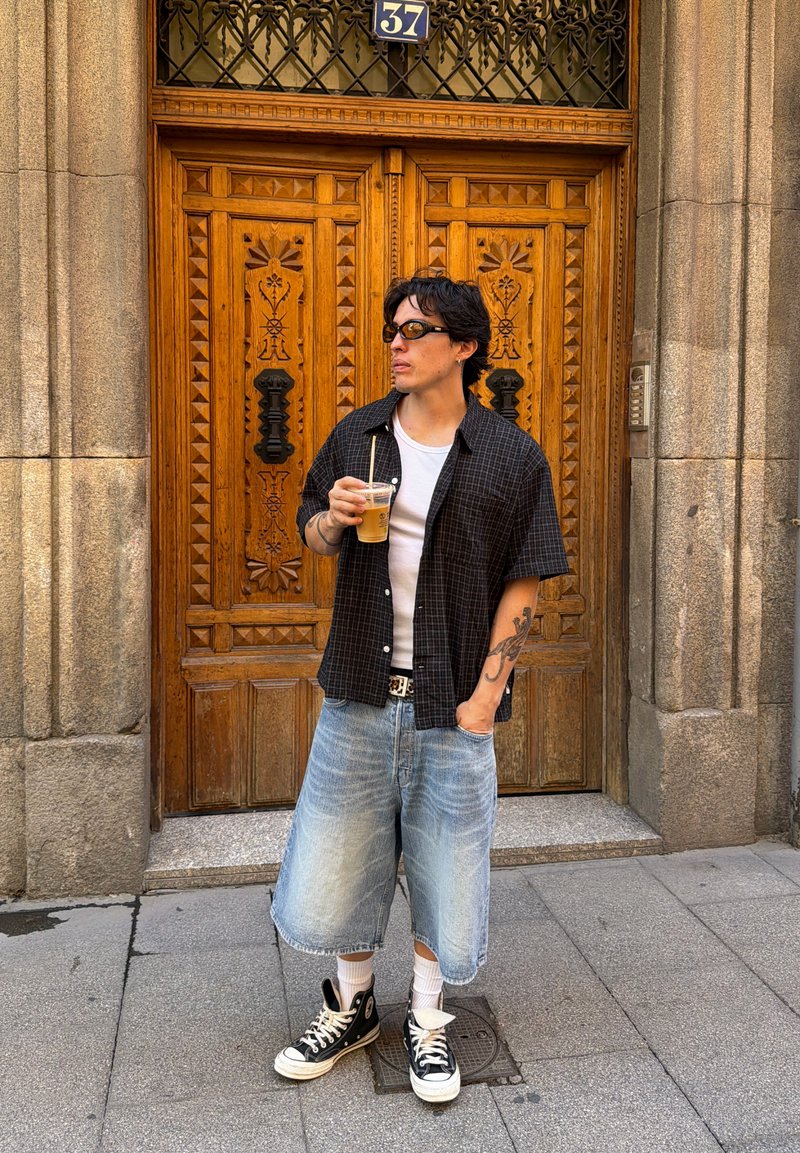 Jeune homme portant des lunettes de soleil, une chemise noire à carreaux et un long short en jean, tenant un café glacé devant des portes doubles en bois finement sculptées.