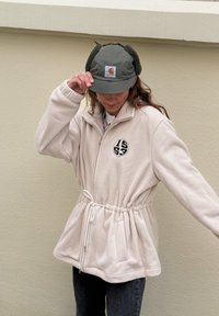 Veste en polaire beige clair avec fermeture à glissière frontale, taille cintrée et logo noir. Associée à une casquette verte avec rabats pour les oreilles et un patch logo.