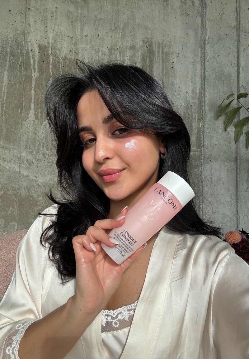 Une femme aux cheveux foncés, vêtue d’un peignoir crème, tient un flacon rose de lotion tonique Lancôme près de son visage. De la crème est appliquée sur sa joue.