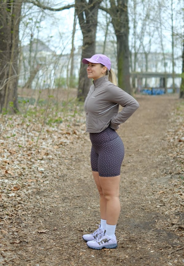 Femeie îmbrăcată în echipament sportiv și șapcă mov stând pe o potecă forestieră cu mâinile în șolduri, înconjurată de copaci și frunze uscate.