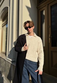 Πλεκτή κρεμ ζακέτα, μαύρο παλτό και τζην παντελόνι. Η ζακέτα διαθέτει V-λαιμόκοψη και ριμπ μανσέτες. Τα γυαλιά ηλίου ολοκληρώνουν την εμφάνιση.