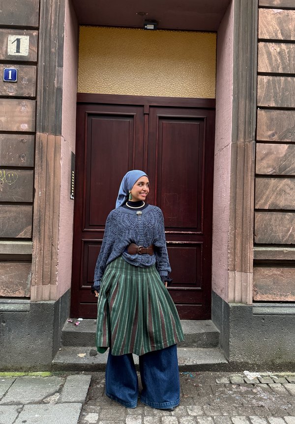 Junge Frau mit blauem Kopftuch, gemusterter blauem Pullover, grün gestreiftem Rock und weiten Jeans steht vor einer Holztür.