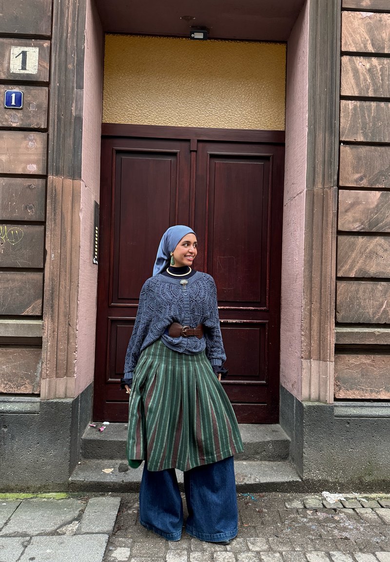 Jeune femme portant un foulard bleu, un pull bleu à motifs, une jupe verte rayée et un jean à jambes larges, debout devant une porte en bois.
