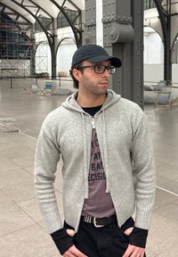 Grå tröja med ribbad nederkant och manschetter, dragkedja fram, bärs över en mörk skjorta med text. Personen har på sig glasögon och en svart mössa. Bakgrunden innehåller arkitektur.