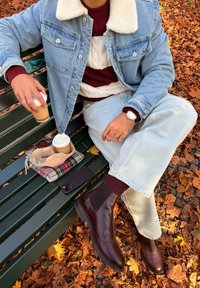 Person i denimjakke, stripete genser, lyse bukser og brune støvler som sitter på en grønn benk med kaffekopper, skjerf og telefon blant høstløv.