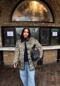 Femme en manteau imprimé léopard, tenant un sac à main noir, se tenant dans une rue pavée devant un mur en briques avec une fenêtre en arc et un comptoir de service.