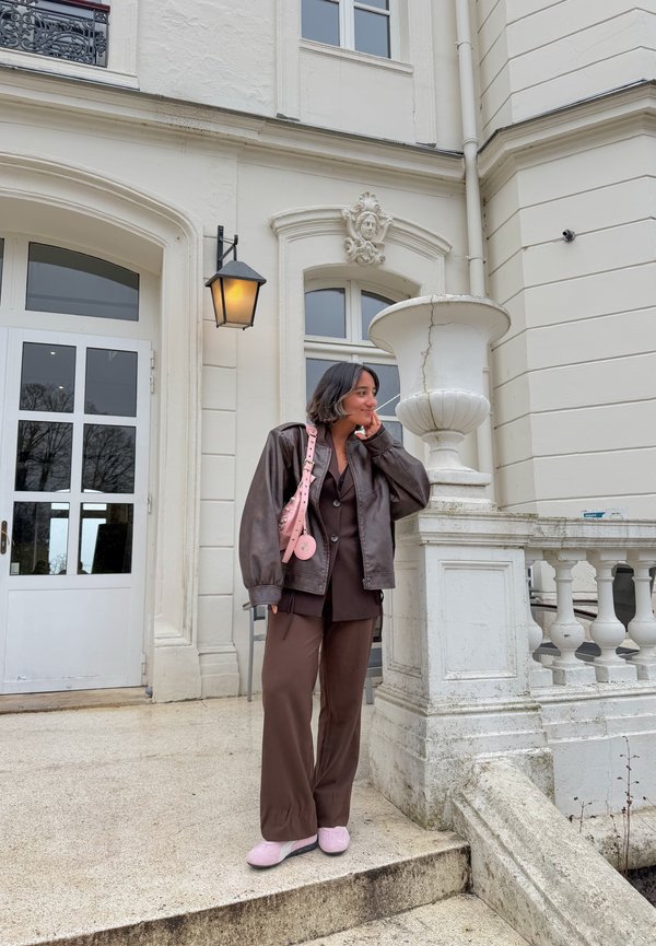 Jeune femme en costume marron et veste en cuir, sac et chaussures roses, debout à côté d'un bâtiment blanc orné avec un grand vase et une lampe.