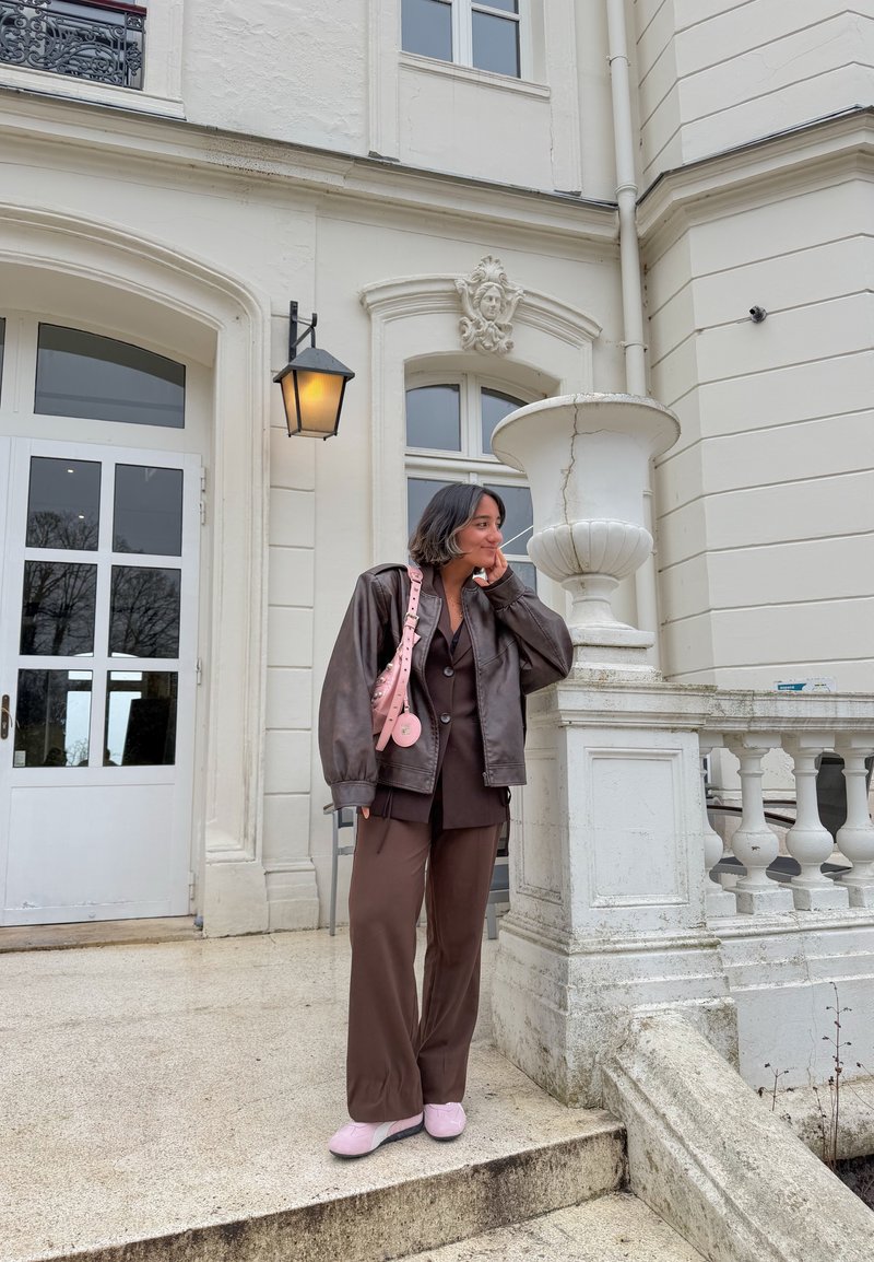 Young woman in brown suit and leather jacket, pink bag and shoes, standing by ornate white building with large urn and lamp.