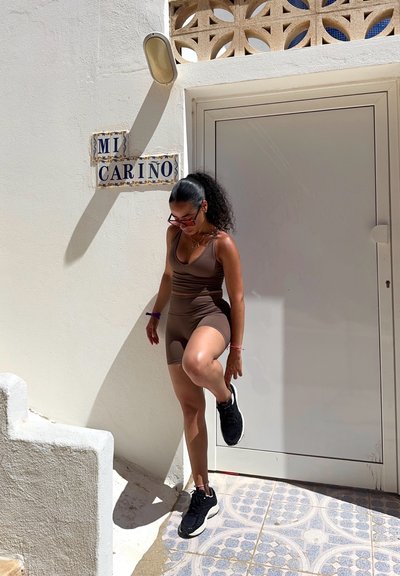 Top de tirantes ajustado marrón y pantalones cortos, zapatillas negras, de pie sobre un mosaico decorado. Pared clara, puerta detrás, cartel decorativo que dice "MI CARINO."