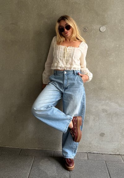 Top corto blanco con encaje, jeans de pierna ancha azul claro y zapatos de plataforma marrones con acentos dorados. Las gafas de sol completan el look.