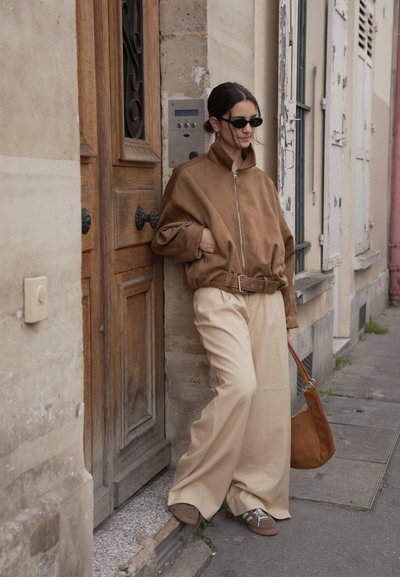 Mujer con chaqueta marrón extragrande y pantalones anchos crema apoyada en el marco de una puerta en una acera de la ciudad, sosteniendo un bolso marrón a juego.