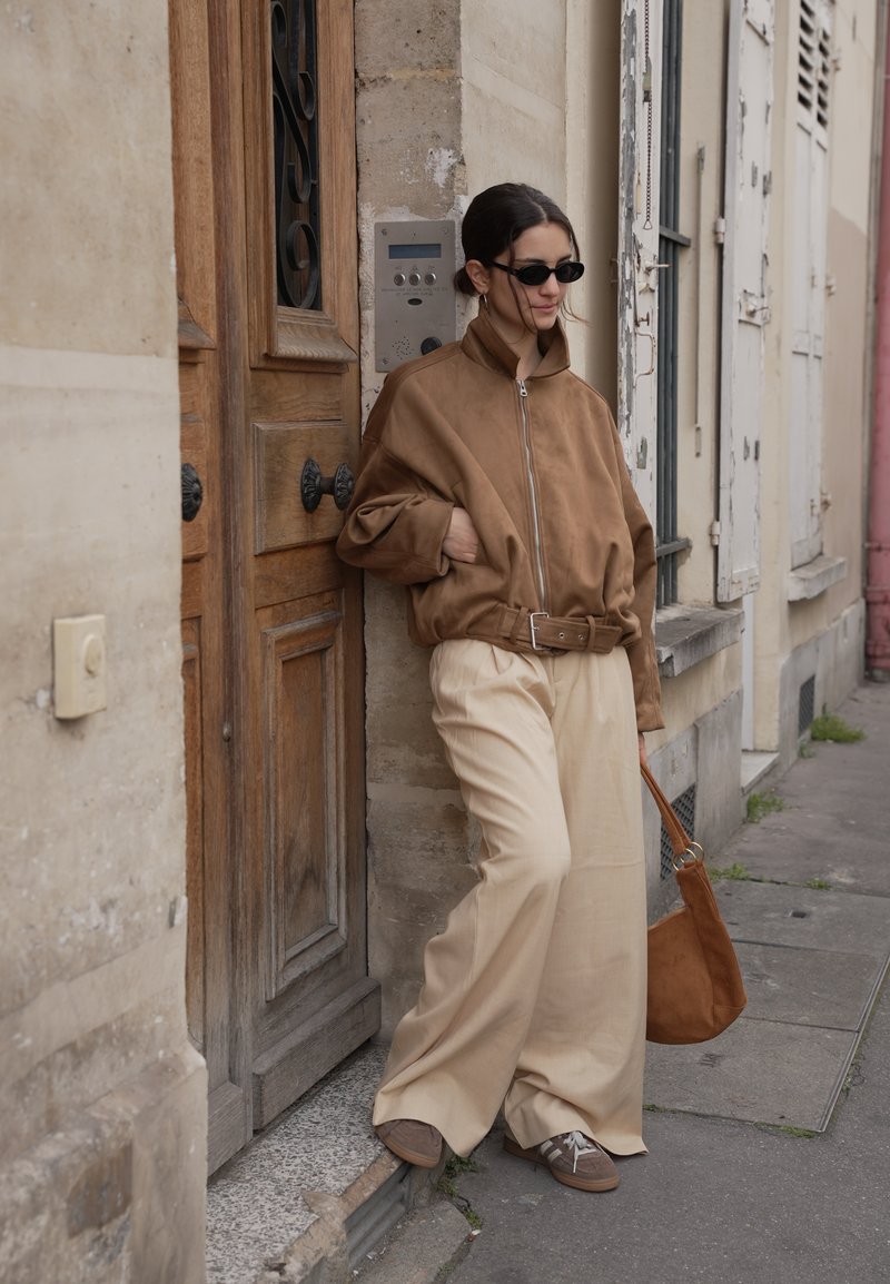 Femme portant une veste marron surdimensionnée et un pantalon large crème, appuyée contre un encadrement de porte sur un trottoir de la ville, tenant un sac à main marron assorti.