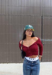 Czerwony sweter o krótkim kroju, dzianinowy, z prążkowaną teksturą, niebieskie dżinsy z wysokim stanem, biała gumka z logo DKNY, zielona czapka baseballowa, okrągłe okulary.