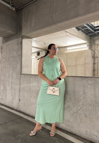 Vestido sin mangas de color verde menta, con botones, textura suave, hasta la rodilla. Combinado con un bolso blanco y sandalias plateadas. Fondo de concreto.