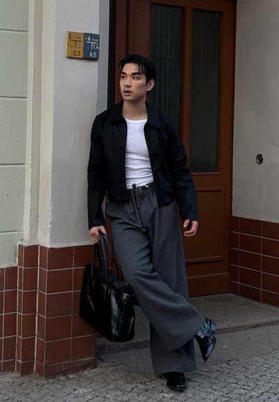 Chaqueta negra corta sobre una camisa blanca, pantalones de pierna ancha gris, llevando una tote negra. Texturas suaves con un ajuste casual. Puerta marrón y pared de azulejos.