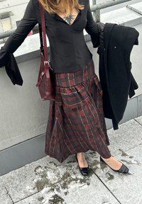 Bluză neagră cu mâneci lungi și manșete evazate, fustă în carouri burgundy cu straturi, balerini negri și geantă burgundy, pe un acoperiș gri.