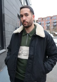 Homme aux cheveux courts et foncés avec une barbe, portant une veste noire avec un col en polaire blanche et un pull vert à fermeture éclair, debout en plein air dans une zone urbaine.