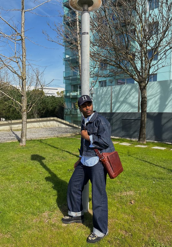 Mann in Jeansjacke und Mütze steht auf Gras, lehnt an einem Laternenpfahl in der Nähe von blattlosen Bäumen und einem modernen Glasgebäude.