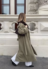 Femme marchant devant un mur de pierre orné, portant un long manteau olive, un pantalon blanc, des chaussures noires pointues, et une sacoche sombre en osier sur l'épaule.