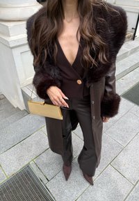 Smeđi kožni kaput s kragnom od umjetnog krzna, uparen s topom dubokog V-izreza, širokim hlačama i zlatnom torbicom. Istaknuti su teksturirani detalji.