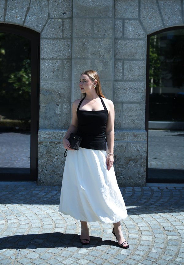 Žena u crnom majici bez rukava i bijeloj suknji stoji na kaldrmi ispred sive kamene zidne površine s dva visoka prozora.