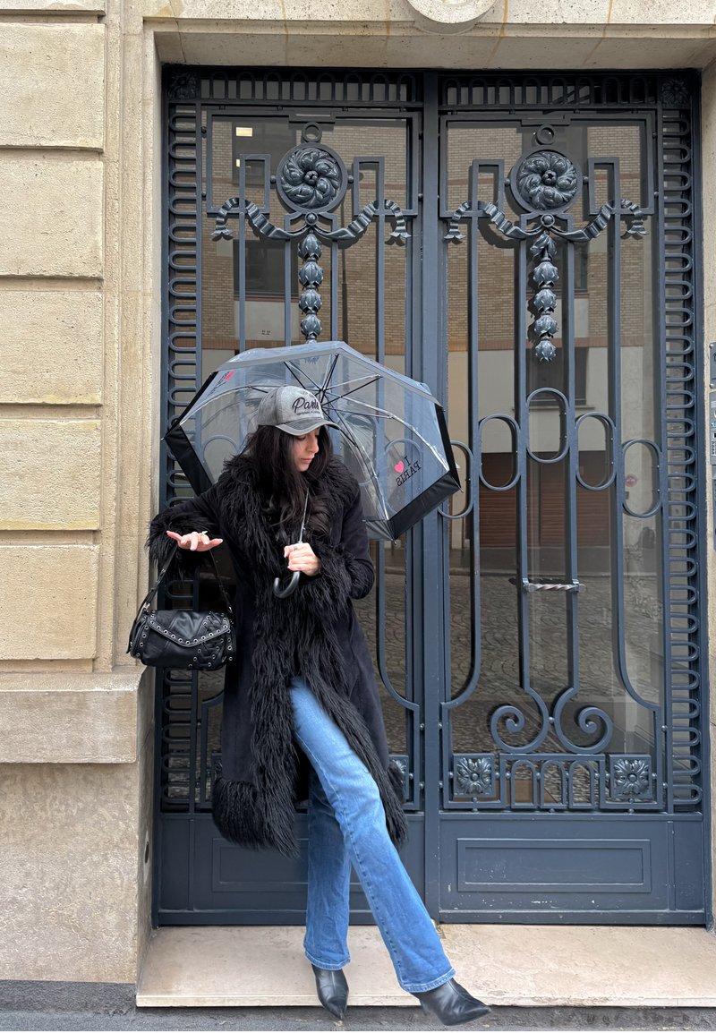 Schwarzer Pelzmantel mit strukturierter Oberfläche, blaue Schlagjeans, hält einen durchsichtigen Regenschirm mit schwarzen Akzenten und eine schwarze Handtasche mit Nieten.
