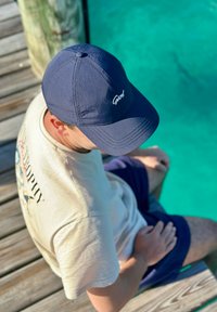Homme portant une casquette bleu marine et une chemise beige, assis sur une jetée en bois, regardant l'eau turquoise claire en dessous.