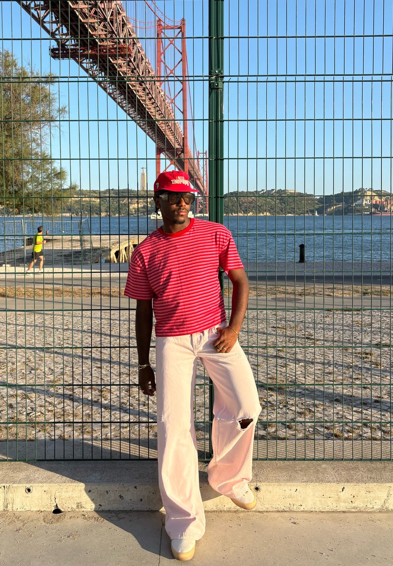 Homme portant une chemise rouge à rayures, un pantalon blanc déchiré et une casquette rouge, debout près d'une clôture verte à côté d'une rivière avec un grand pont rouge en arrière-plan.