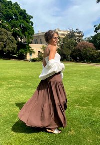 Kobieta w okularach przeciwsłonecznych, ubrana w białą bluzkę, brązową, zwiewną spódnicę oraz białe sandały, spacerująca po zielonym trawniku w pobliżu drzew i beżowego budynku.