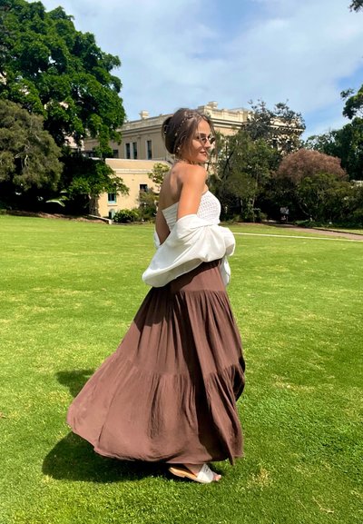Mujer con gafas de sol, camiseta blanca, falda marrón fluida y sandalias blancas caminando sobre césped verde cerca de árboles y un edificio beige.
