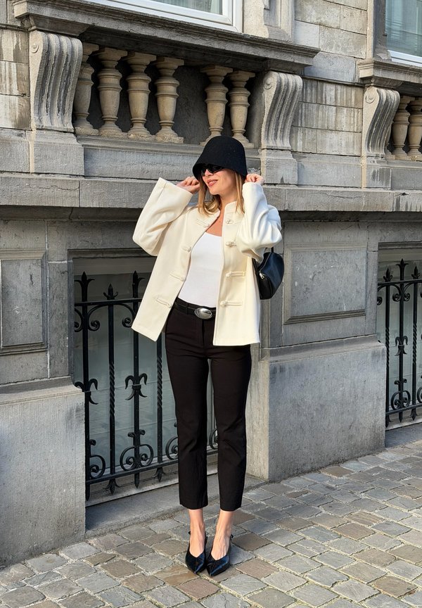 Una mujer con chaqueta blanca, pantalones negros, sombrero y gafas de sol se ajusta el sombrero frente a un edificio histórico de piedra.